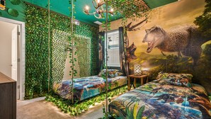 Ein Zimmer mit einer grünen, blätterbedeckten Wand, einem Wandgemälde eines Dinosauriers und einem Bett mit einer farbenfrohen Bettdecke im Dino-Design.