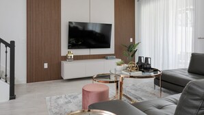 A modern living room with a flat-screen TV mounted on a wall, a glass coffee table, a grey sofa, and a pink ottoman. - Last Minute Disney Getaway! Private Pool & Themed Bedrooms. Book Now! #111841 (Orlando)