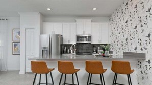 A modern kitchen with white cabinets, a stainless steel refrigerator, a microwave, and a large island with bar stools. - Last Minute Disney Getaway! Private Pool & Themed Bedrooms. Book Now! #111841 (Orlando)