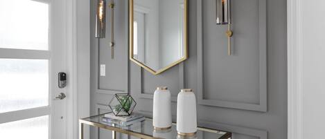 A modern interior with a glass console table, two white vases with gold accents, a hexagonal mirror, and two wall-mounted light fixtures.