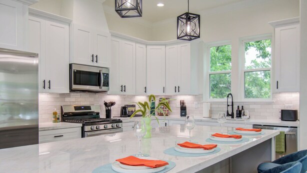 A modern kitchen with white cabinets, a central island with blue placemats, and a large window with a view of trees. - Rooftop Movie Oasis: 3bd - Games - Patio (Houston)