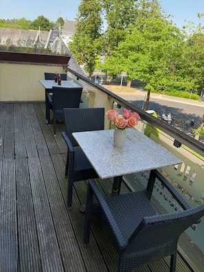 Une terrasse en bois avec des sièges d’extérieur, une table agrémentée d’un vase de fleurs et une vue sur une rue et des arbres.