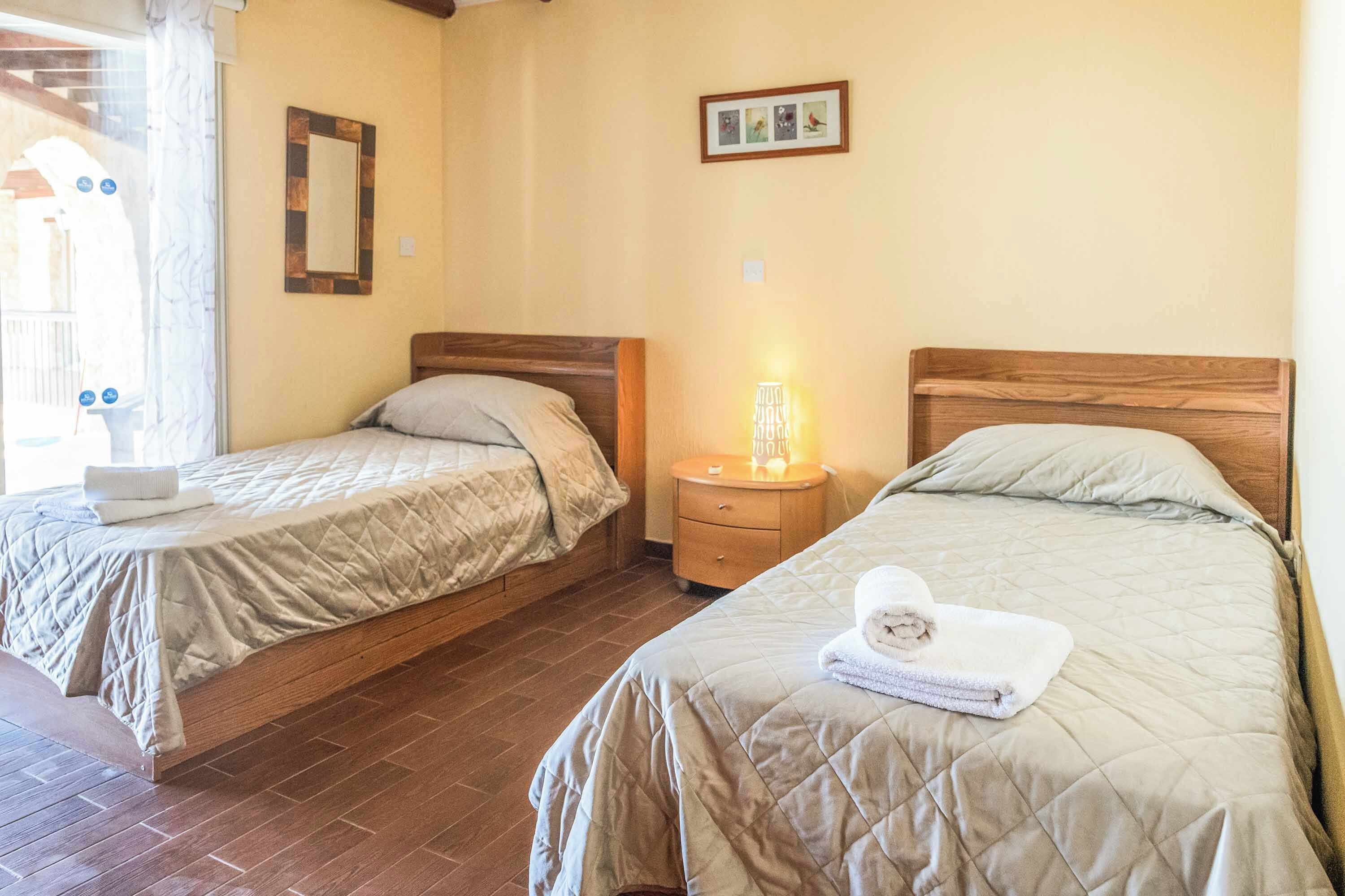 A room with two beds, each with a wooden headboard, a bedside table with a lamp, and a framed picture on the wall.