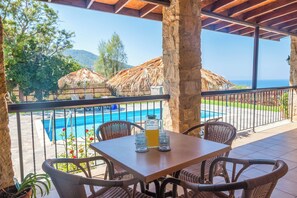 A covered outdoor seating area with a table and chairs, overlooking a swimming pool and a view of the sea.