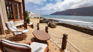 A beachside terrace with a wooden table and two lounge chairs, overlooking a rocky shoreline and the sea.