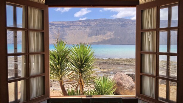 A room with a view of a beach and mountains through a window with curtains.