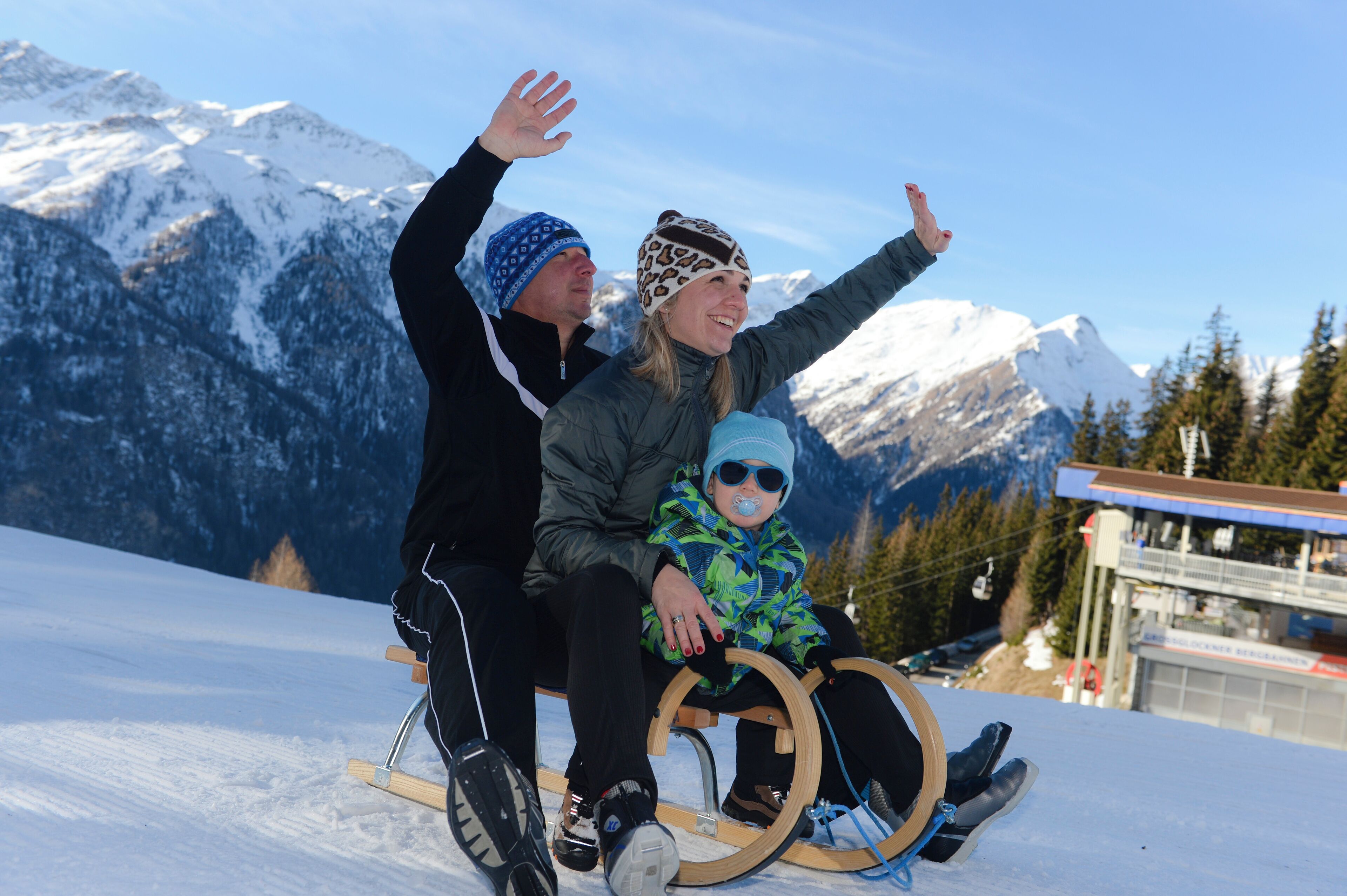 Eine Person und ein Kind auf einem Schlitten an einem verschneiten Hang, im Hintergrund Berge.