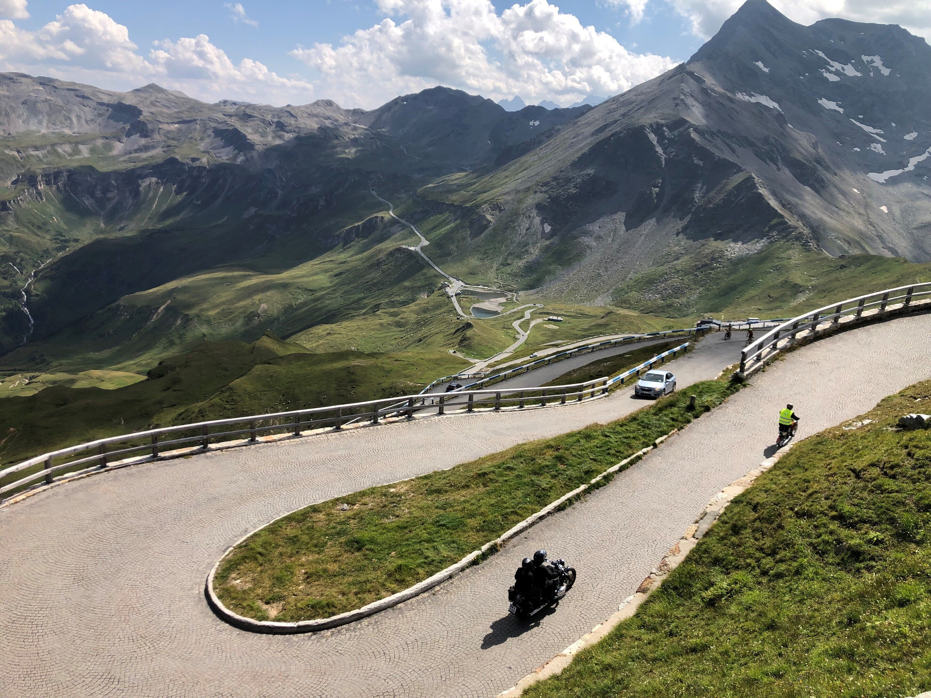 A winding mountain road with a guardrail, surrounded by lush green hills and distant peaks.