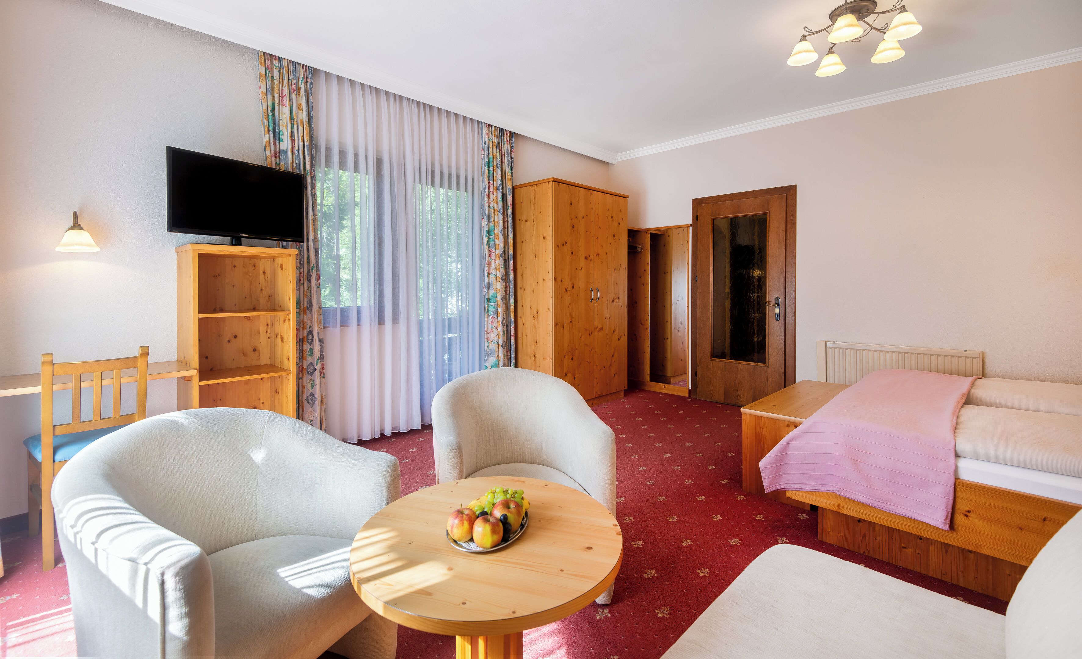 Une chambre d’hôtel avec un lit, une armoire en bois, une télévision, un bureau et une table ronde sur laquelle sont disposés des fruits.