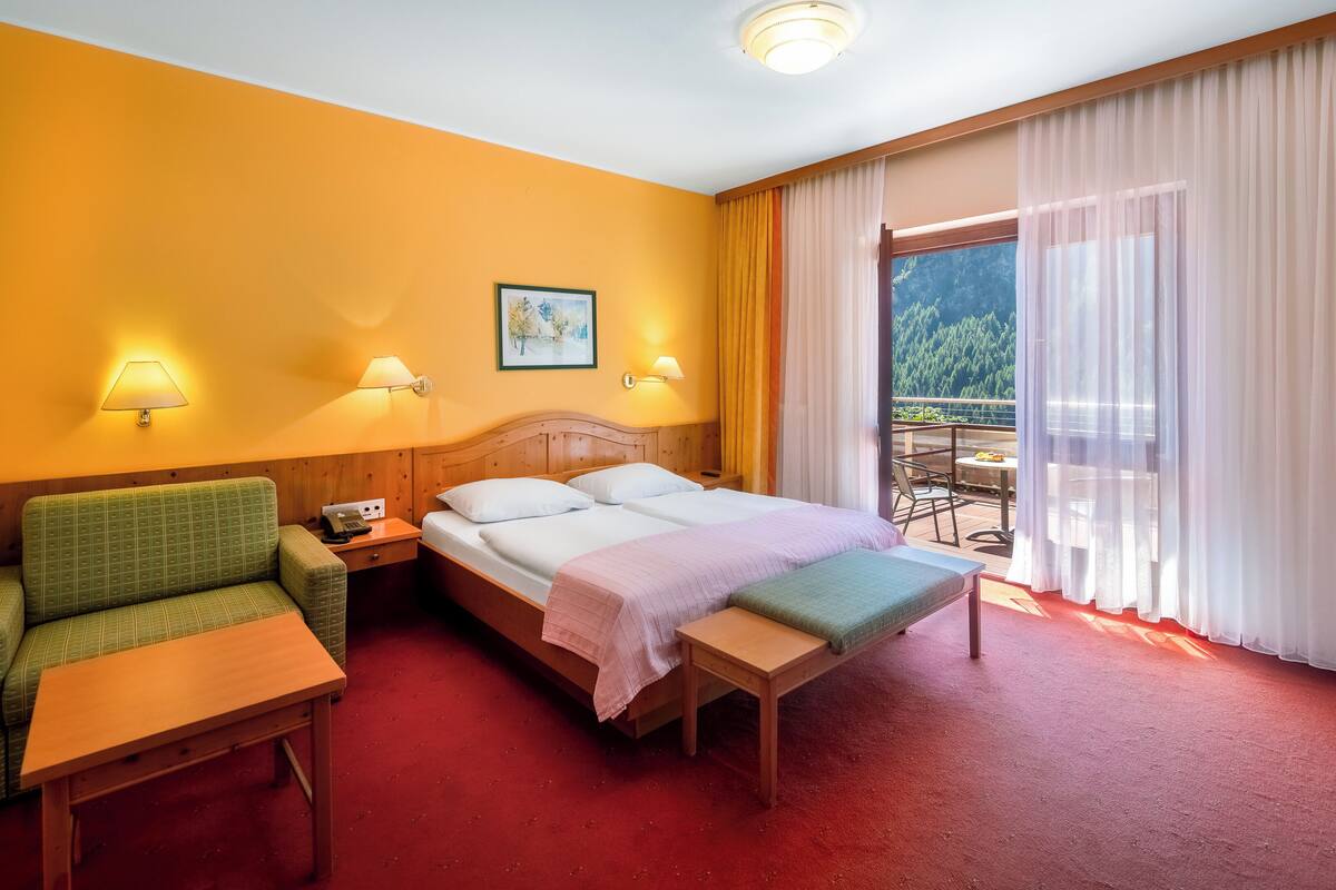 Una habitación de hotel con una cama, un sofá verde, una mesita de noche de madera, una mesa y un balcón con vistas a árboles.