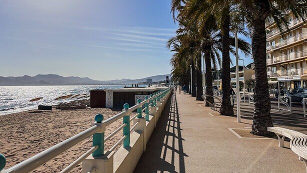 A coastal promenade with a beach, palm trees, and a row of buildings with balconies. - Apartment 3 bedrooms 3 min to beach (Cannes)