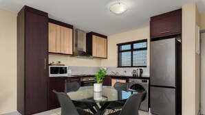 A modern kitchen with dark wooden cabinets, a stainless steel refrigerator, a microwave, and a glass dining table with four chairs. - Modern Century City Apartment - Mountain Views (Cape Town)