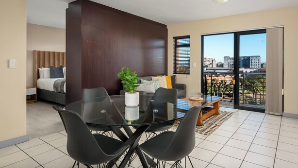 A modern living room with a glass dining table, black chairs, a sofa, and a large window offering a city view. - Modern Century City Apartment - Mountain Views (Cape Town)
