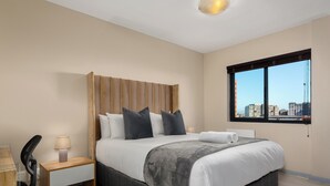 A modern hotel room with a large bed, a wooden headboard, a desk with a chair, and a window with a city view. - Modern Century City Apartment - Mountain Views (Cape Town)