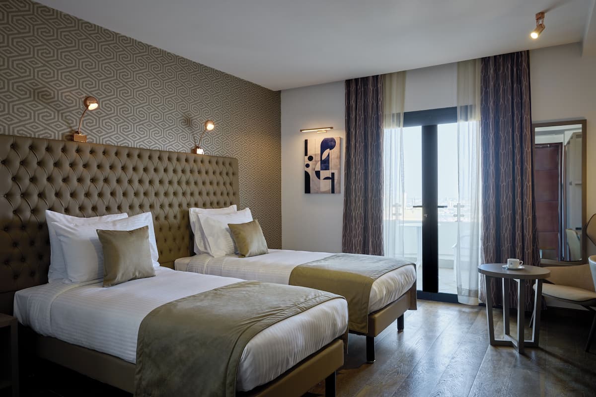 Hotellihuone, jossa on kaksi sänkyä, koristeltu pääty, kuvioitu seinä, taulu ja näkymä ulos ovesta.