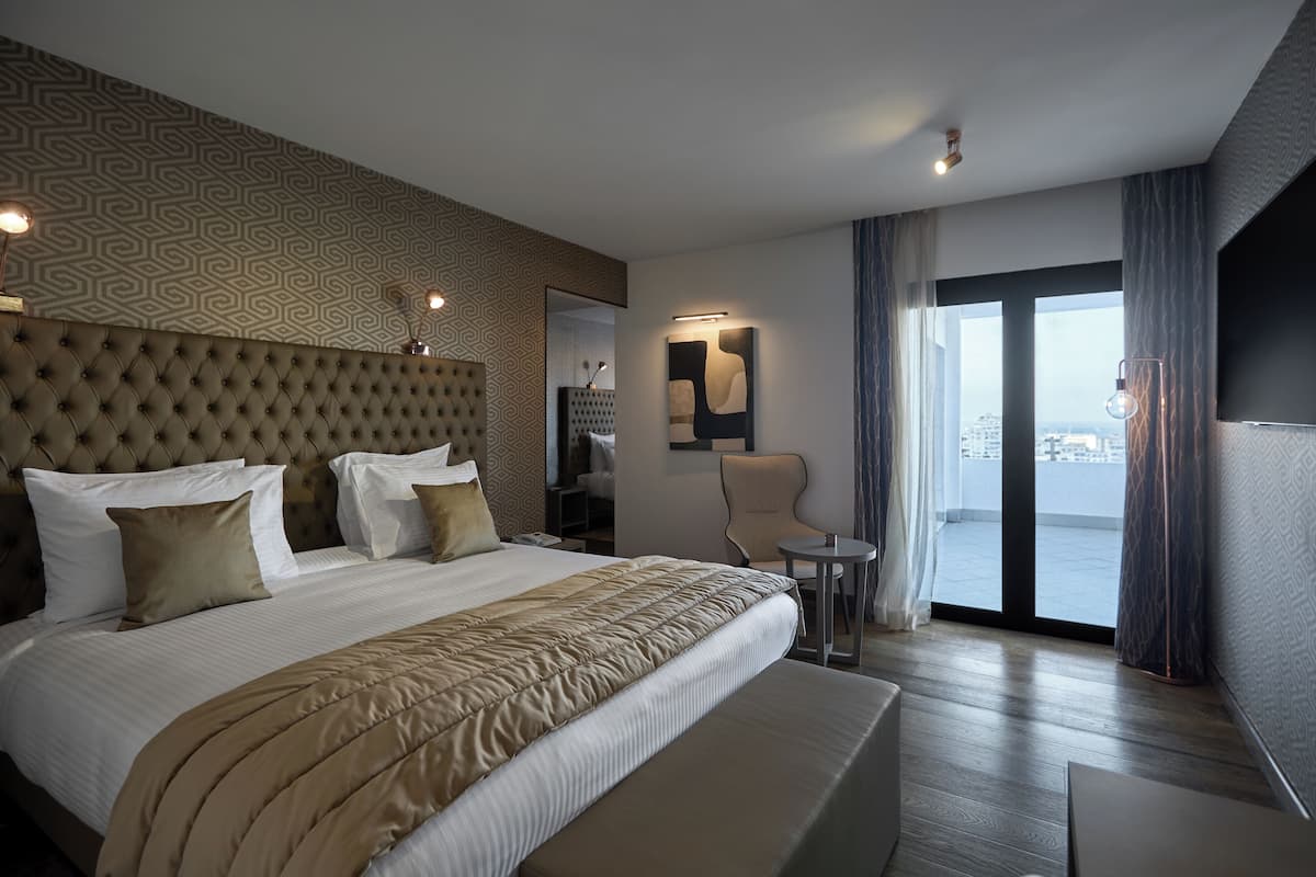 Moderni hotellihuone, jossa on suuri sänky, oleskelualue ja näkymä lasiovien läpi.