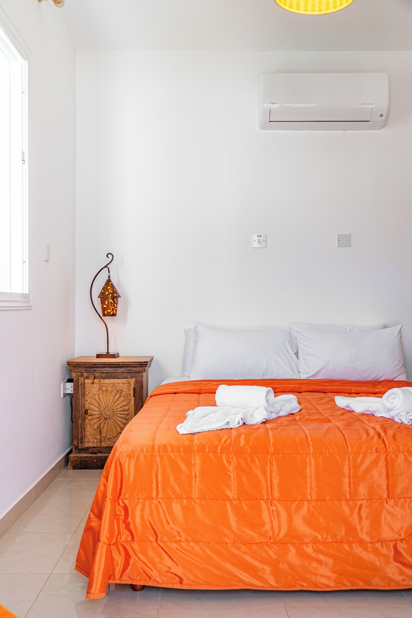 A bedroom with a bed, white pillows, an orange bedspread, a wooden nightstand with a lantern, and an air conditioning unit on the wall.