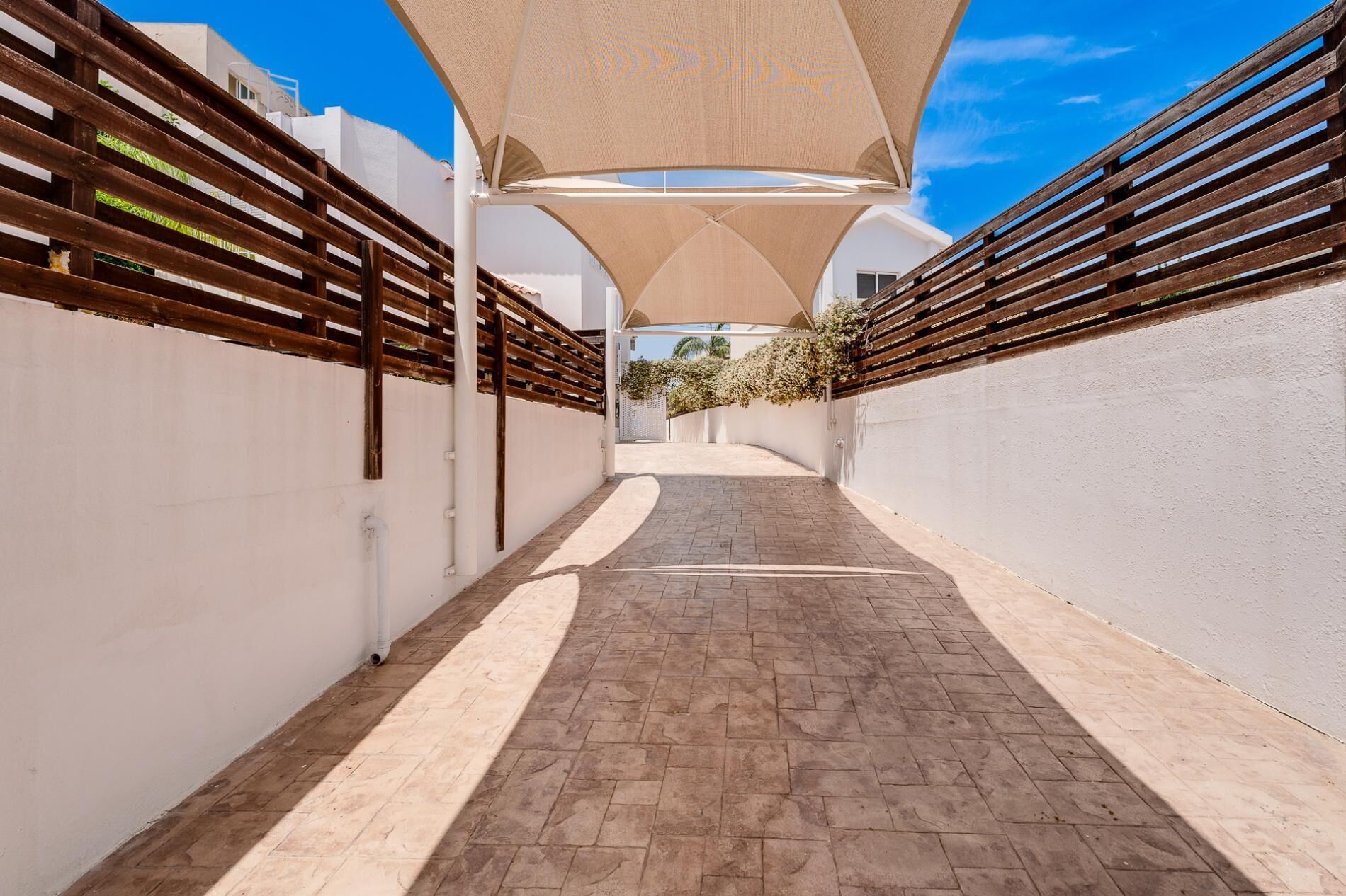 A narrow pathway with a brick floor, flanked by white walls and wooden fences, leading to a building with a white facade and a large canopy structure.
