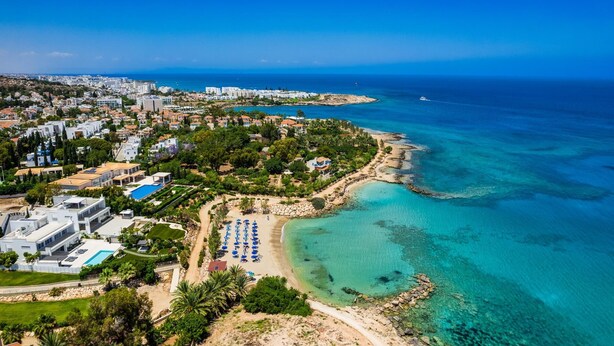 An aerial view of a coastal resort with a beach, clear turquoise waters, and a mix of residential and hotel buildings.