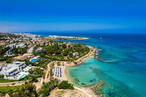 Vue aérienne d’une station balnéaire avec une plage, des eaux turquoise et limpides, et un mélange de bâtiments résidentiels et d’hôtels.