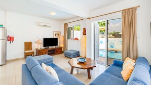 A living room with a TV, sofa, and a small table. There is a sliding glass door leading to an outdoor area with a pool.