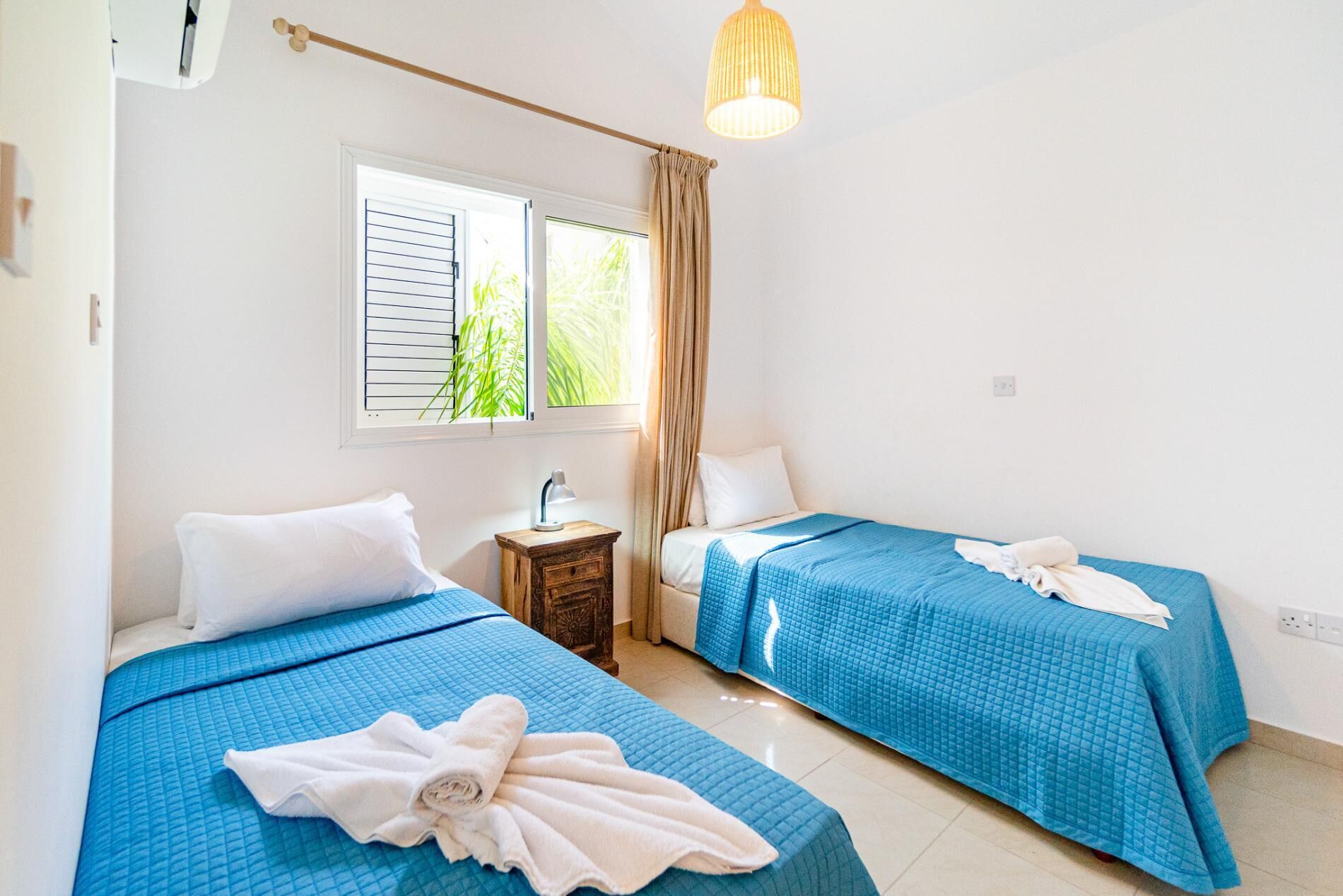 Habitación de hotel con dos camas, cada una con cubrecama azul acolchado y almohadas blancas. Entre las camas, hay una mesita de noche de madera con una lámpara. Se ve una ventana con persianas y cortina, y un candelabro colgante sobre las camas.