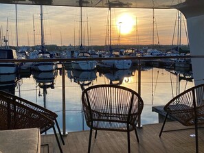 Vista desde la cubierta de un barco al atardecer, con varias sillas y una mesa, con vistas a un puerto deportivo con numerosos barcos.