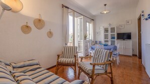Een woonkamer met een bank, twee gestreepte fauteuils, een eettafel met stoelen, een televisie en een keukenhoek met kastjes.