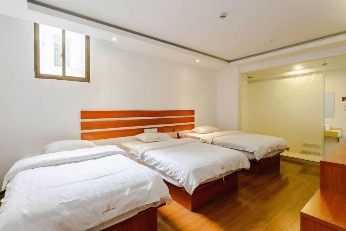Habitación de hotel con tres camas, cabecera de madera, un ventanal grande y zona de ducha con mampara de vidrio.