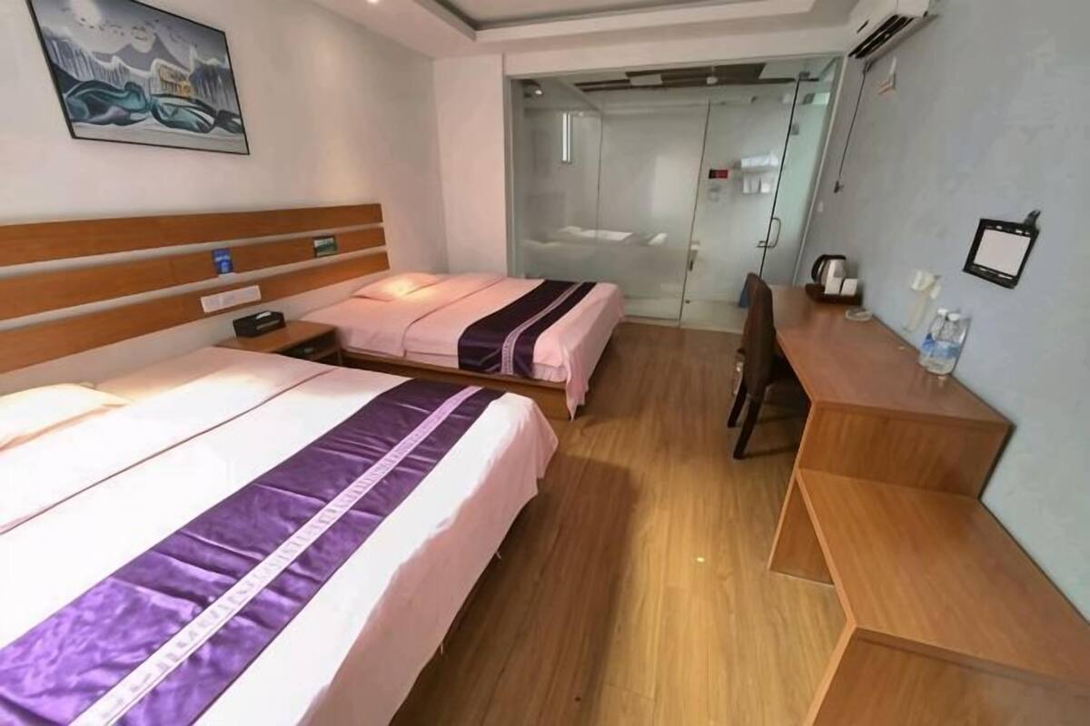 Habitación de hotel con dos camas, un escritorio de madera, una silla, una mesita y una puerta de cristal que da al baño.