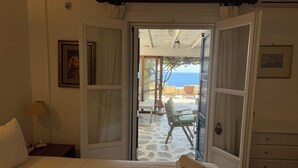 A hotel room with a bed, a dresser, a chair, and a view of the sea through an open door. - Elia Oasis 2 Bd Magic Villa w Sea Views (Mykonos)