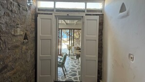 A stone-walled room with a wooden ceiling, a white door leading to a stone-floored area with a table and chairs, and a view of the outdoors. - Elia Oasis 2 Bd Magic Villa w Sea Views (Mykonos)