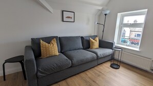 A grey sofa with patterned cushions, a small black side table, a floor lamp, and a window with a view of buildings. - Top Floor Apartment (Llandudno)
