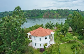 Une maison blanche à deux étages, surmontée d’un toit recouvert de tuiles rouges, est entourée d’une végétation luxuriante et bordée par une rivière.