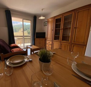 Ein Essbereich mit Holztisch, gedeckt für zwei Personen, einer Zimmerpflanze und Blick auf eine gebirgige Landschaft durch ein großes Fenster.