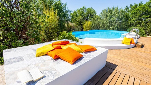 A poolside area with a wooden deck, a white outdoor seating area with cushions, and a clear blue pool. - Villa Ambra by Villa Plus (San Vito dei Normanni)