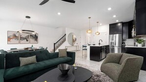A modern living room with a green sofa, a round coffee table, and a gray armchair. There is a kitchen area with black cabinets and stainless steel appliances.