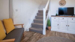 Una sala de estar con un sofá gris, un banco de madera, una escalera, un televisor sobre un mueble bajo y una planta en maceta.