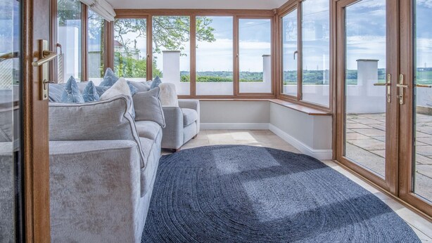 Une pièce moderne avec un canapé gris, un tapis bleu et de grandes fenêtres donnant sur un paysage verdoyant et une vue lointaine.