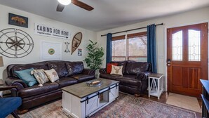 A furnished living room with two leather sofas, a coffee table, a ceiling fan, a large window with curtains, and a door with glass panels. - Stroll to Park, Shops, Restaurants in Downtown WJ! (West Jefferson)
