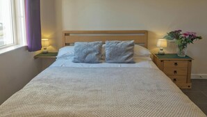 A bed with a wooden headboard, two pillows, a white blanket, a nightstand with a lamp, and a vase with flowers. - Llainfawr Cottage Apartment, Fishguard (Letterston)