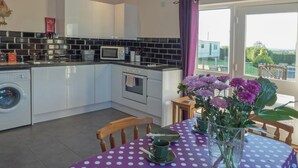A modern kitchen with a washing machine, microwave, and oven. There is a dining area with a table set for two and a view of the outdoors. - Llainfawr Cottage Apartment, Fishguard (Letterston)