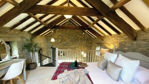 Un dormitorio con pared de piedra, vigas de madera, una cama con ropa de cama blanca, un escritorio con silla y una escalera.