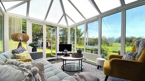 Un invernadero con sofá, sillón y mesa de centro, una televisión y vistas a un paisaje verde.