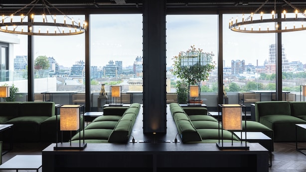 A modern lounge area with green sofas, tables, and lamps, featuring large windows with a city view.