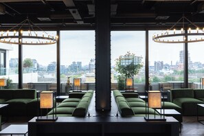 A modern lounge area with green sofas, tables, and lamps, featuring large windows with a city view.
