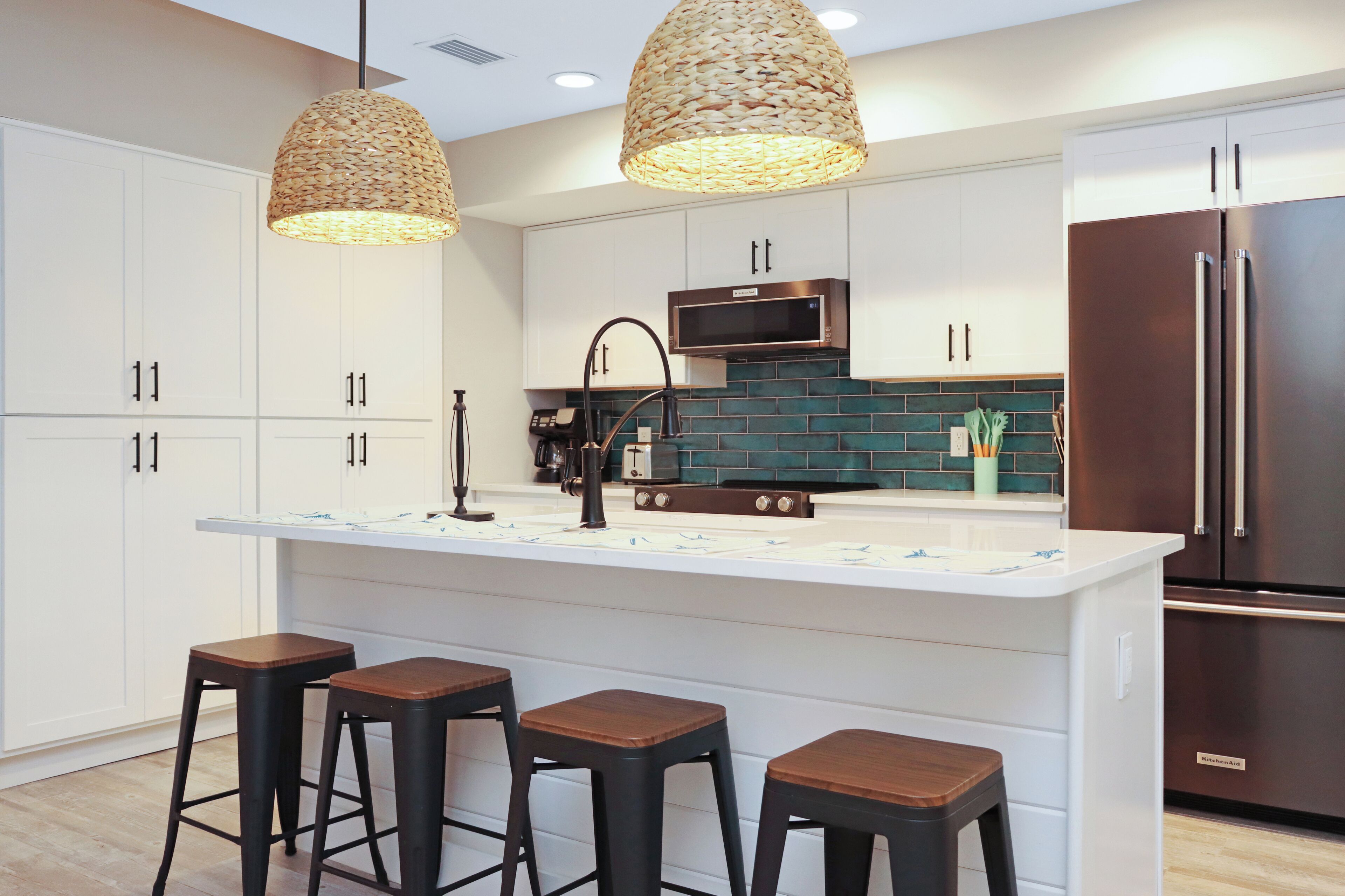 A modern kitchen with white cabinets, a central island with stools, a stainless steel refrigerator, and a woven pendant light.
