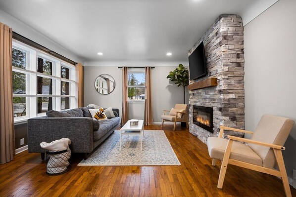 A modern living room with a stone fireplace, a grey sofa, a coffee table, and a large window with curtains.