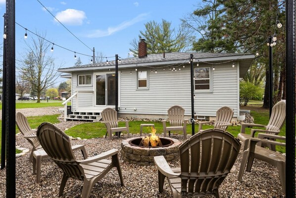 A cozy outdoor seating area with Adirondack chairs and a fire pit, set against a white house with a chimney. - NEW Bellaire Home, 5 Minute WALK to downtown! (Bellaire)