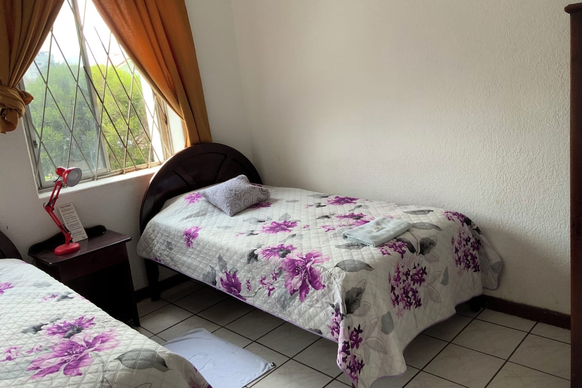 Un dormitorio con una cama individual, una mesita de noche, una lámpara de escritorio y una ventana con cortinas.